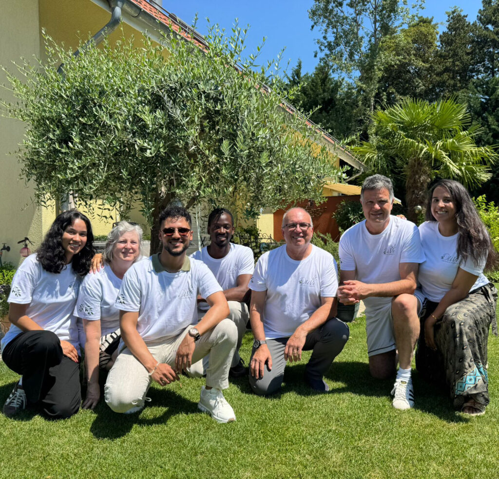 Un groupe de sept membres de l'équipe de l'Association OliVe, portant des t-shirts blancs assortis avec le logo, posant ensemble pour une photo en plein air sur une pelouse ensoleillée. En arrière-plan se trouvent un grand olivier, un palmier et une maison jaune.