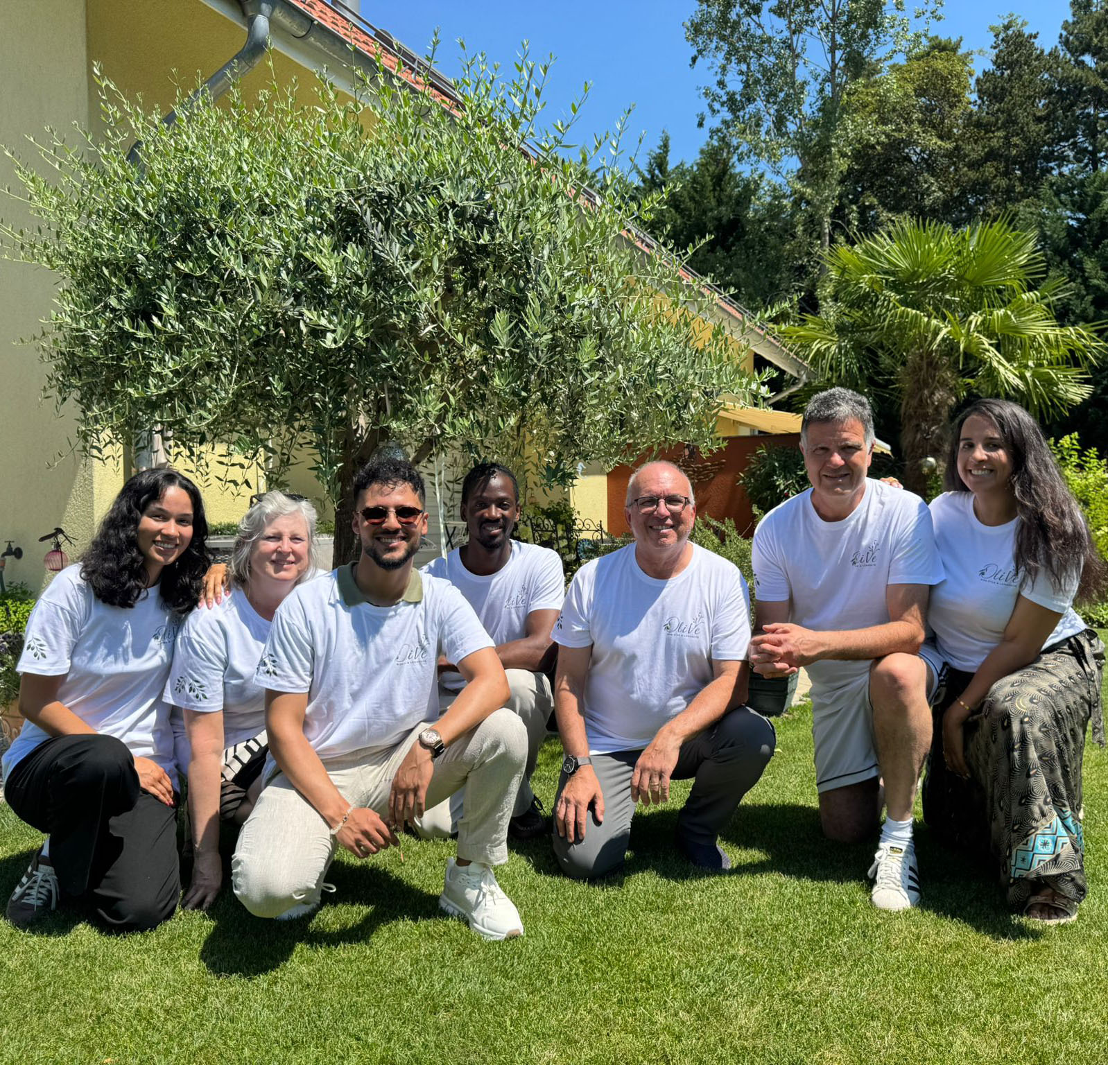Un groupe de sept membres de l'équipe de l'Association OliVe, portant des t-shirts blancs assortis avec le logo, posant ensemble pour une photo en plein air sur une pelouse ensoleillée. En arrière-plan se trouvent un grand olivier, un palmier et une maison jaune.