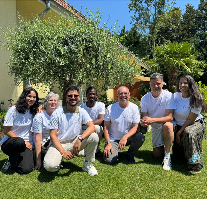 Un groupe de sept membres de l'équipe de l'Association OliVe, portant des t-shirts blancs assortis avec le logo, posant ensemble pour une photo en plein air sur une pelouse ensoleillée. En arrière-plan se trouvent un grand olivier, un palmier et une maison jaune.