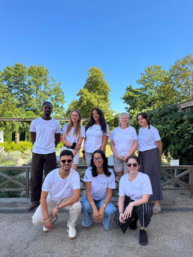 Huit membres de l'équipe de l'Association OliVe, vêtus de t-shirts blancs au logo de l'association, posant ensemble en extérieur dans un parc arboré lors d'une journée ensoleillée.