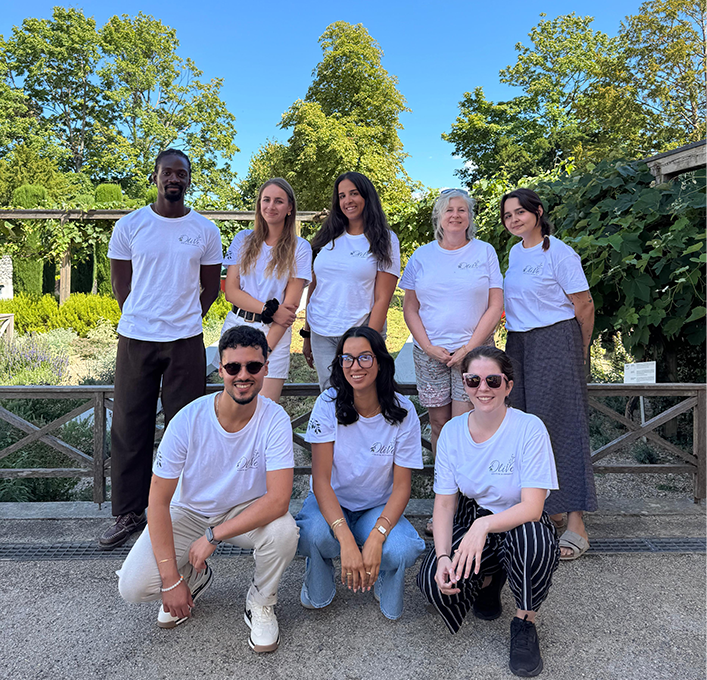 Huit membres de l'équipe de l'Association OliVe, vêtus de t-shirts blancs au logo de l'association, posant ensemble en extérieur dans un parc arboré lors d'une journée ensoleillée.