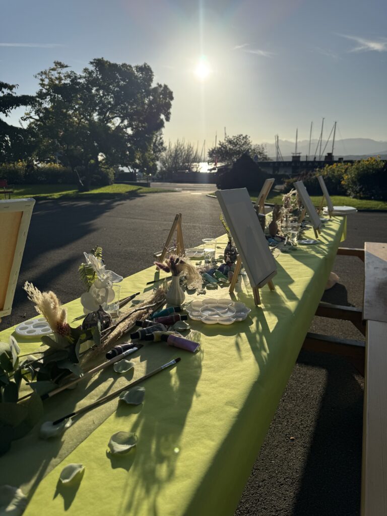 La table de l’atelier en plein soleil, avec chevalets et fleurs séchées, le lac en arrière-plan.