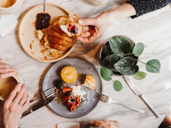 Vue de dessus d’une table de café : croissants, assiettes garnies, tasses et mains qui se servent.